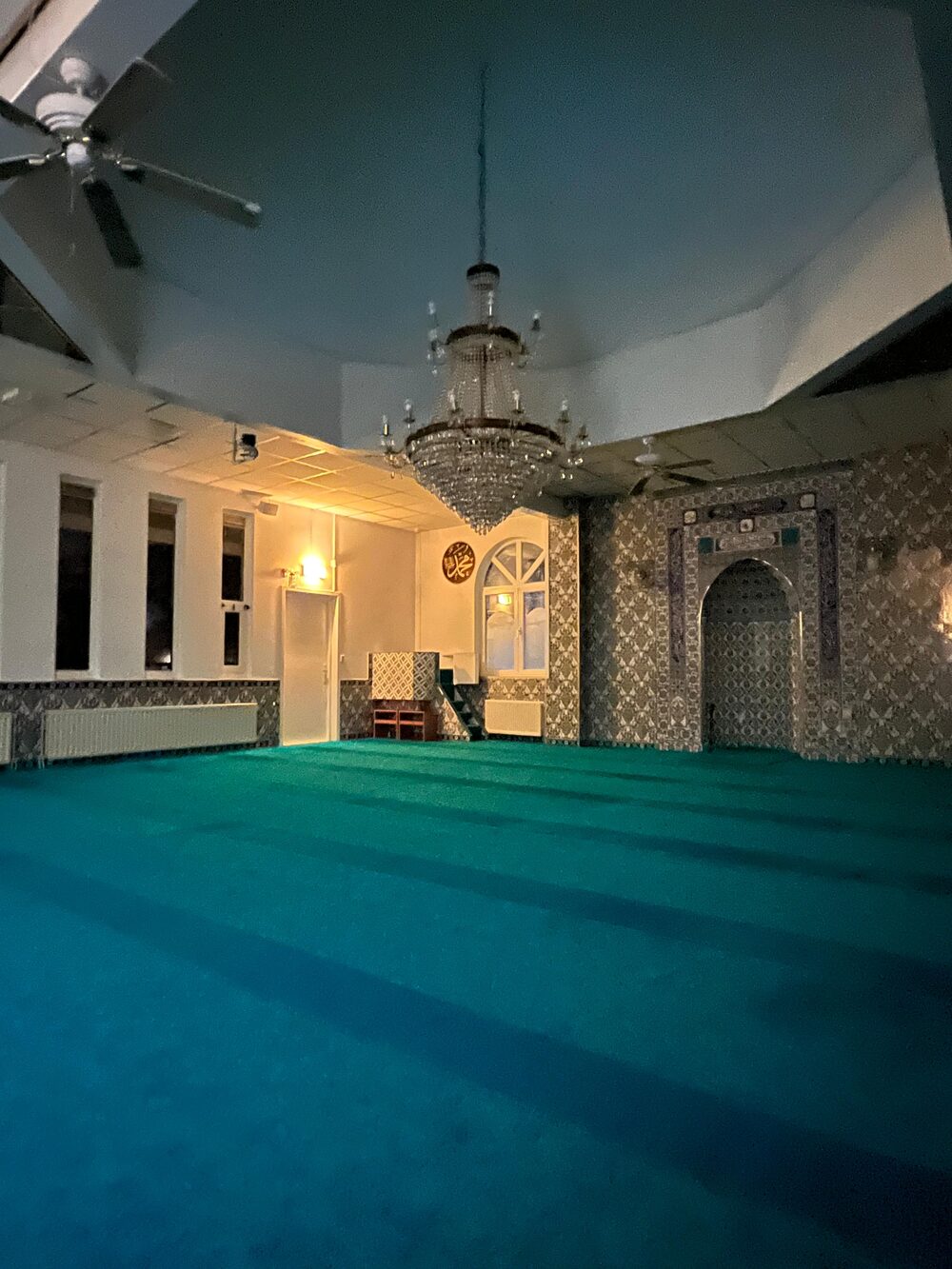 De gebedsruimte in de avond met het turquoise tapijt, de kroonluchter en de mihrab